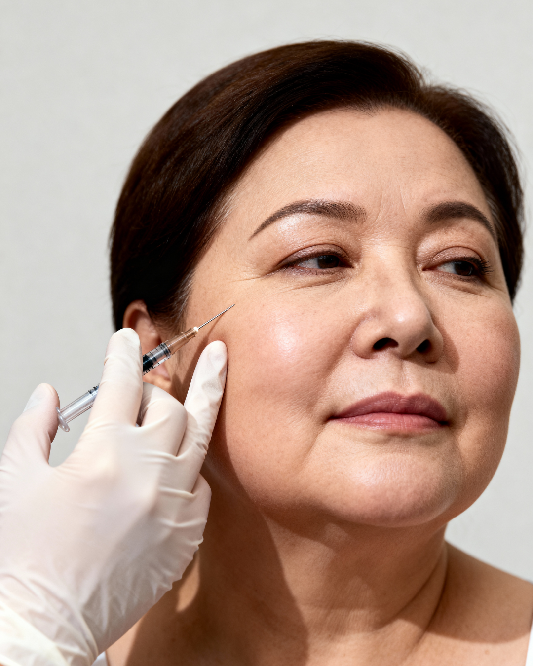 Close-up of a 45-50 year old woman with a fuller face receiving a dermal filler injection in her cheekbone area. A specialist's gloved hand holds the syringe. Illustrates the procedure for Ozempic Face correction